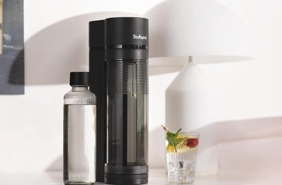 Schwarzer Wassersprudler mit Glasflasche auf einem Tisch neben einem gefüllten Wasserglas