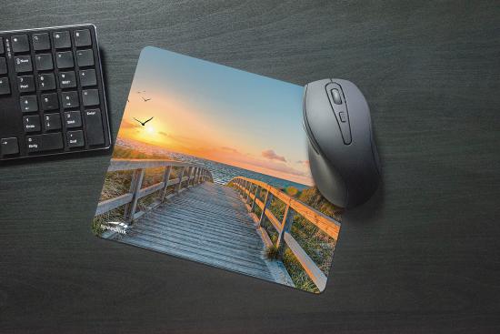 Ein Mauspad mit einem Bild eines Holzstegs zum Strand bei Sonnenuntergang, daneben eine Computermaus auf einem dunklen Schreibtisch.