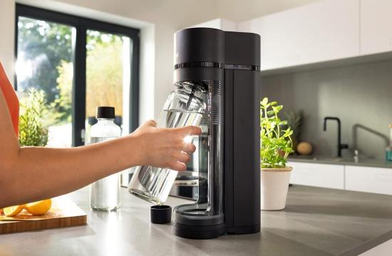 Eine Person füllt eine Glasflasche mit Wasser an einem schwarzen Wassersprudler in einer modernen Küche.