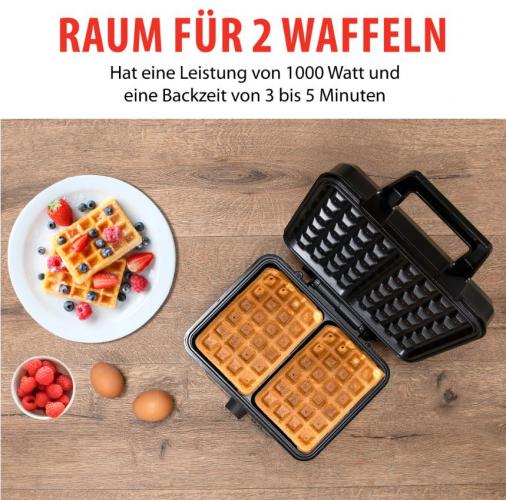 Ein Waffeleisen mit zwei gebackenen Waffeln neben einem Teller mit frischen Waffeln und Beeren sowie Eiern auf einem Holztisch