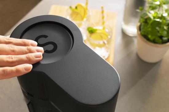 Ein Sodastream wird von einer Hand bedient, im Hintergrund stehen Gläser mit Zitronenwasser und eine Pflanze.