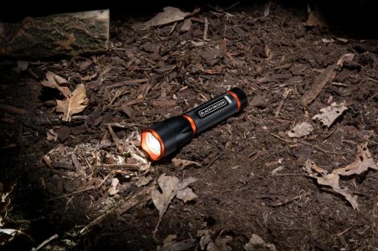 Schwarze und orangefarbene LED-Taschenlampe liegt leuchtend auf Waldboden mit Laub und Zweigen