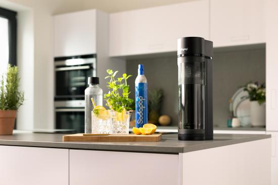 Ein moderner Wassersprudler auf einer Küchenarbeitsplatte mit Gläsern, Zitrone und Kräutern im Hintergrund.