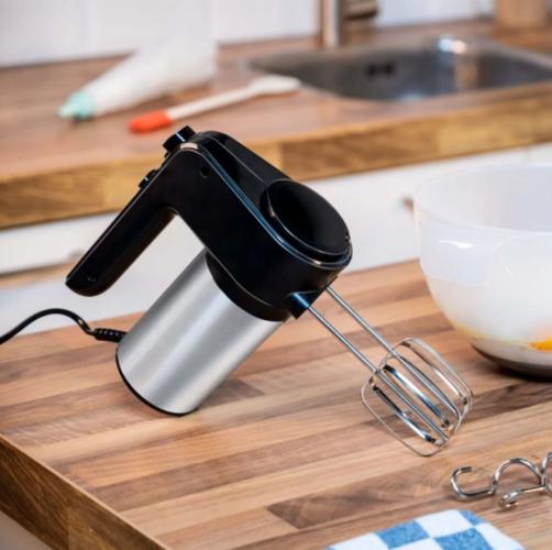 Ein elektrischer Handmixer in einer Küche auf einer Holzarbeitsplatte, neben einer Schüssel und Küchenutensilien platziert.