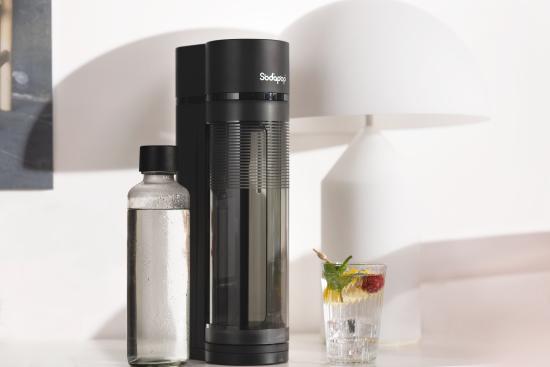 Schwarzer Wassersprudler neben einer Glasflasche und einem Glas mit Zitronenscheiben, Minze und Beeren