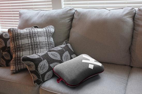 Ein graues Sofa mit mehreren dekorativen Kissen in verschiedenen Mustern vor einem Fenster mit weißen Jalousien