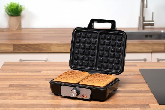 Ein modernes, schwarzes elektrisches Waffeleisen, das zwei frisch gebackene Waffeln zeigt, steht auf einer Holzarbeitsplatte in einer Küche.