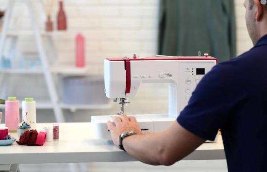 Person bedient eine Nähmaschine auf einem Tisch, umgeben von Garnspulen und Zubehör in einem kreativen Studio.